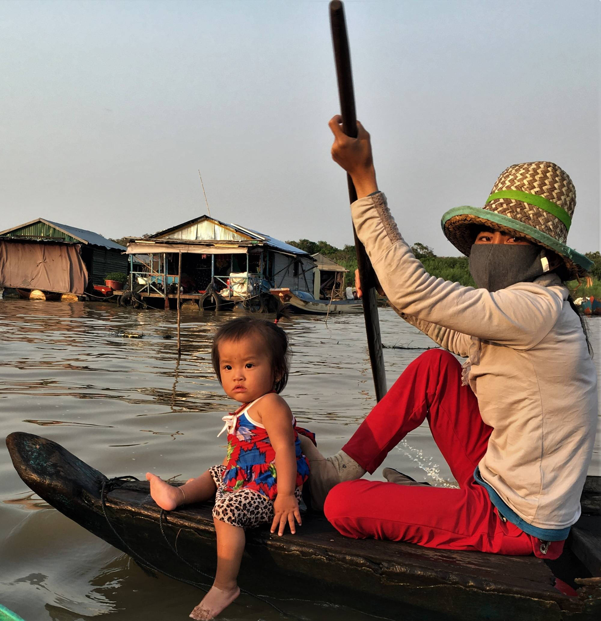 Colored photograph of a woman wearing a mask and a young toddler on a small boat or canoe. Buildings floating in the water behind them